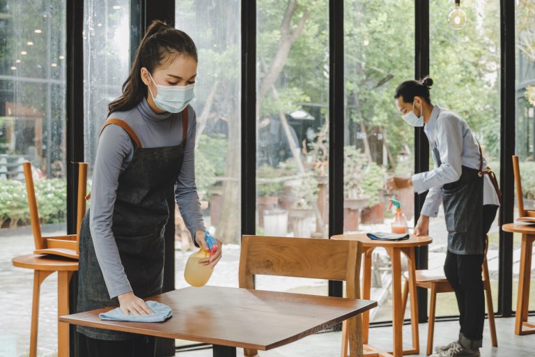 asian waitress staff wearing protection face mask in apron cleaning table with disinfectant spray for protect infection coronavirus (covid-19) in cafe coffee shop restaurant. new normal concept